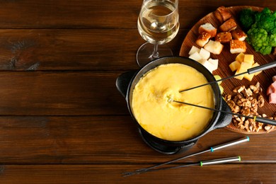 Fondue pot with melted cheese, glass of wine and different products on wooden table, flat lay. Space for text