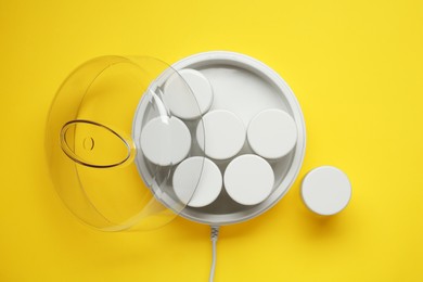 Modern yogurt maker with jars on yellow background, flat lay