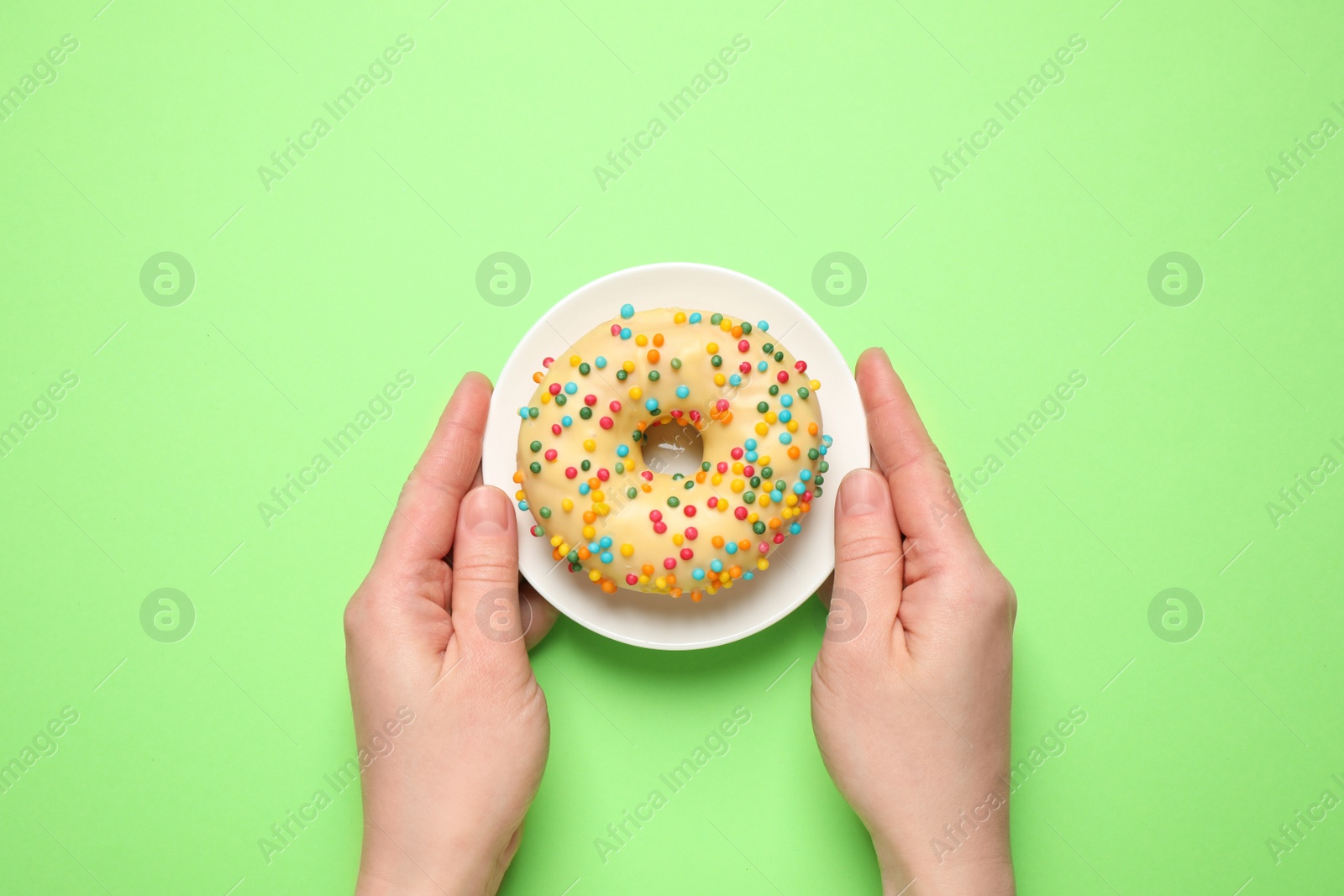 Photo of Woman holding plate with delicious glazed donut on green background, top view