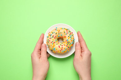 Woman holding plate with delicious glazed donut on green background, top view