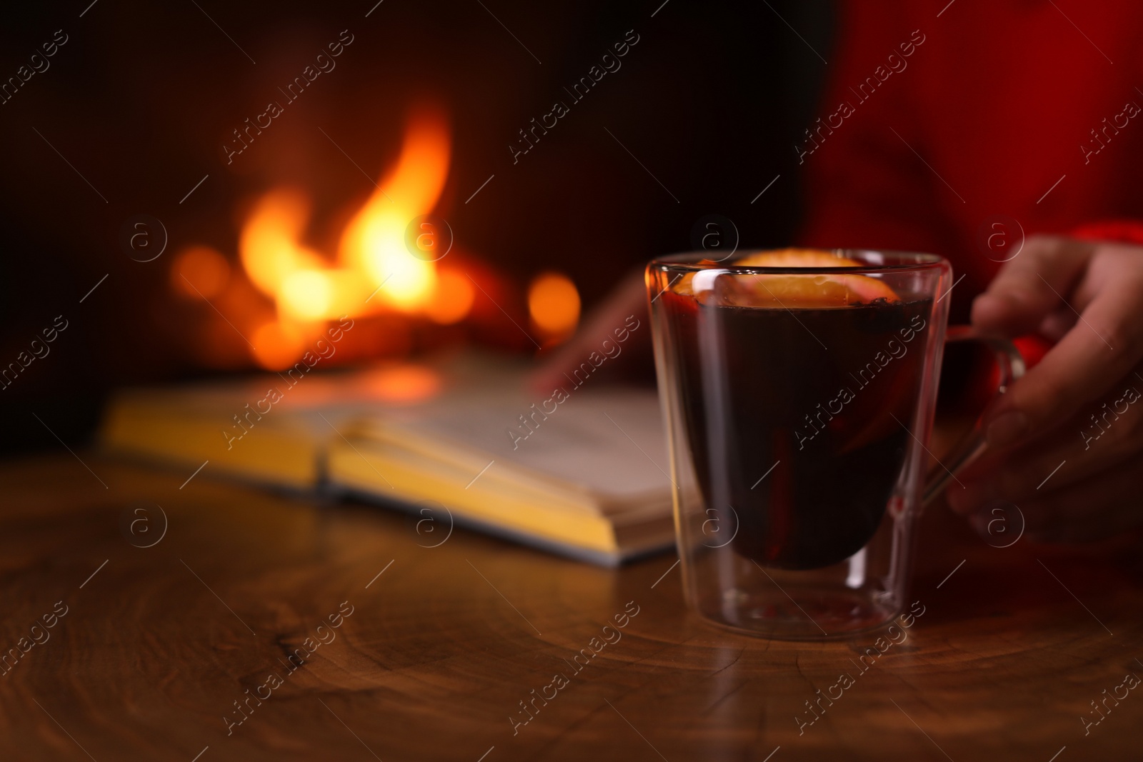Photo of Woman with mulled wine reading book near fireplace indoors, closeup. Winter vacation