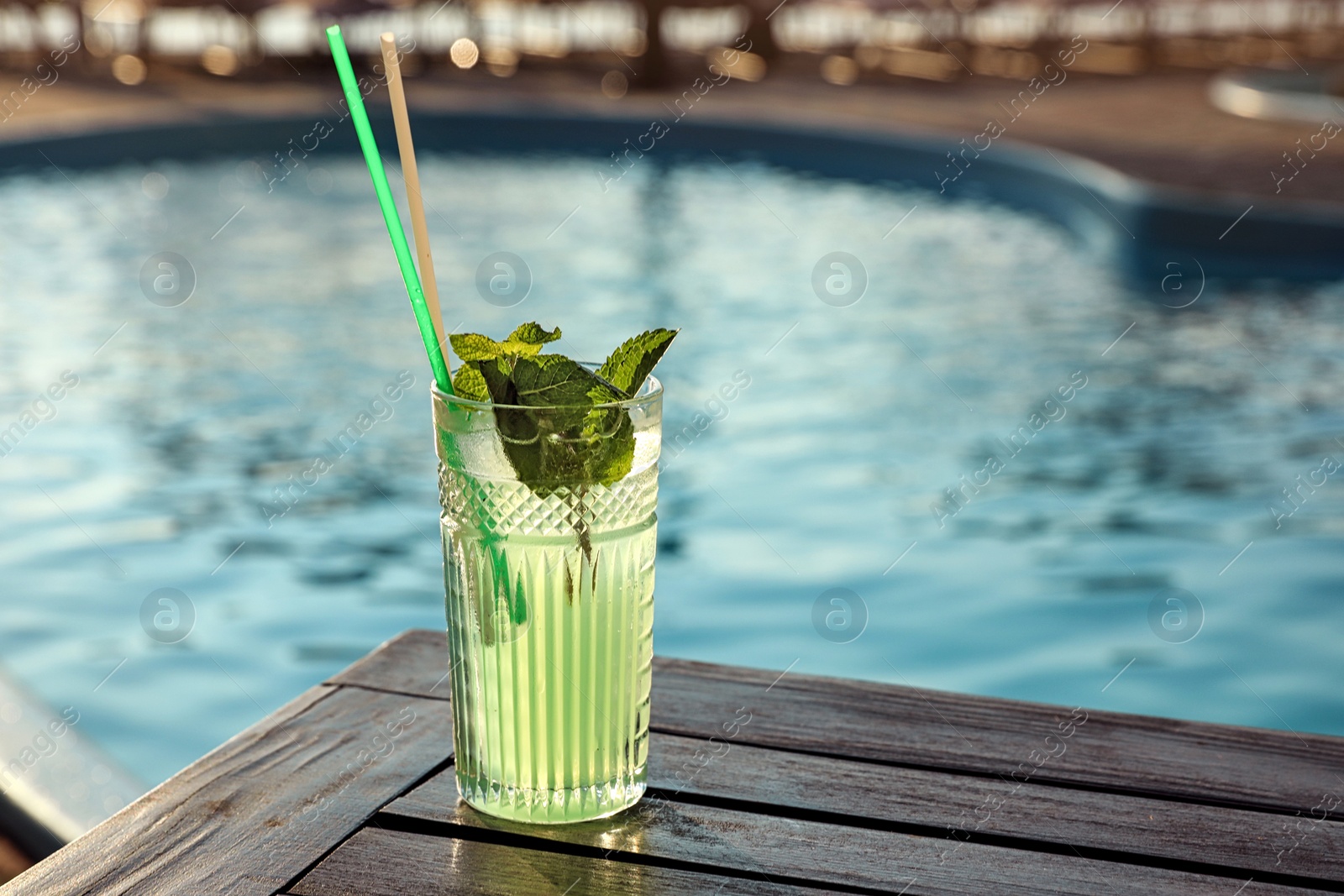 Photo of Glass of fresh summer cocktail on wooden table near swimming pool outdoors. Space for text