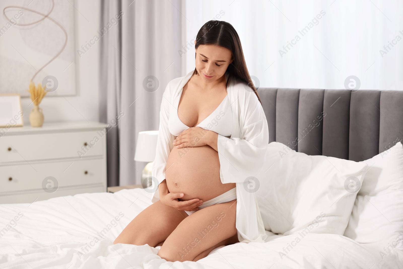 Photo of Beautiful pregnant woman in stylish comfortable underwear and robe on bed at home