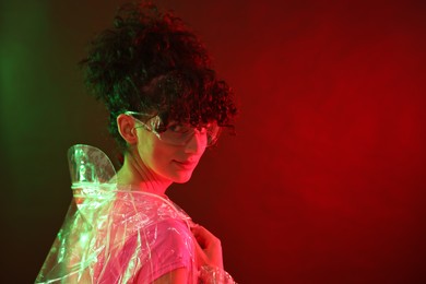 Photo of Beautiful young woman in transparent coat and sunglasses posing on color background in neon lights. Space for text