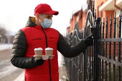 Courier in medical mask holding takeaway drinks and ringing gate bell outdoors. Delivery service during quarantine due to Covid-19 outbreak