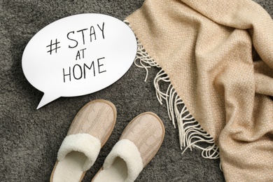 Photo of Slippers, plaid and speech bubble with hashtag STAY AT HOME on grey carpet, flat lay. Message to promote self-isolation during COVID‑19 pandemic