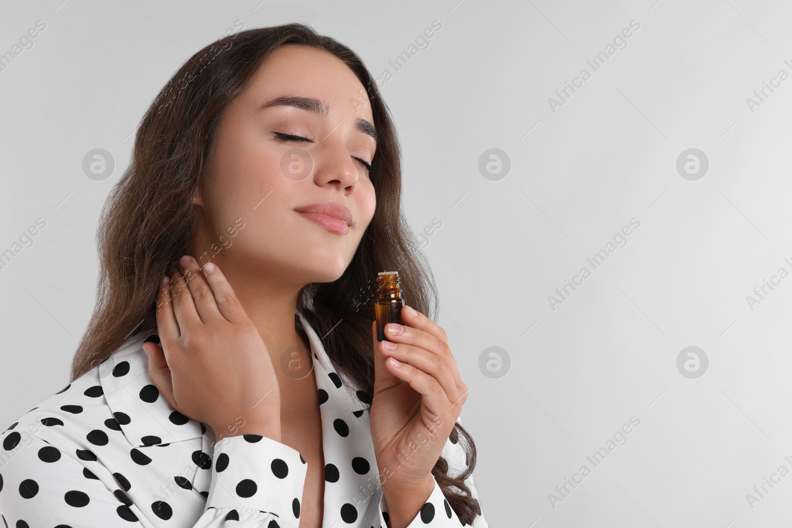 Photo of Beautiful young woman applying essential oil onto neck on light grey background, space for text