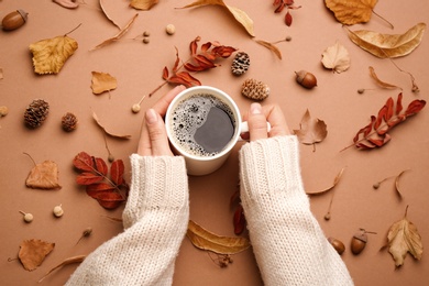 Photo of Woman with cup of hot drink surrounded by autumn leaves on brown background, flat lay. Cozy atmosphere