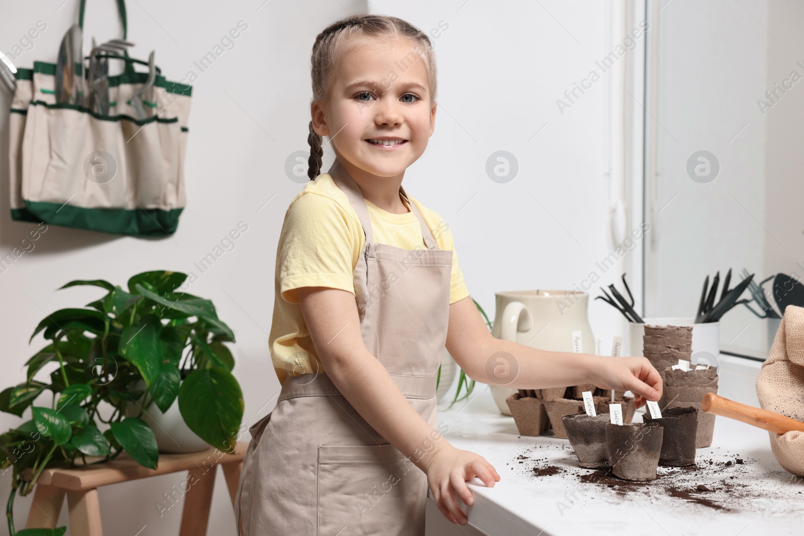 Photo of Little girl inserting cards with names of vegetable seeds into peat pots on window sill indoors