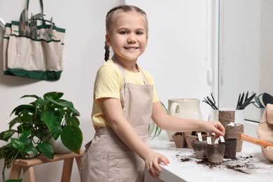 Photo of Little girl inserting cards with namesvegetable seeds into peat pots on window sill indoors