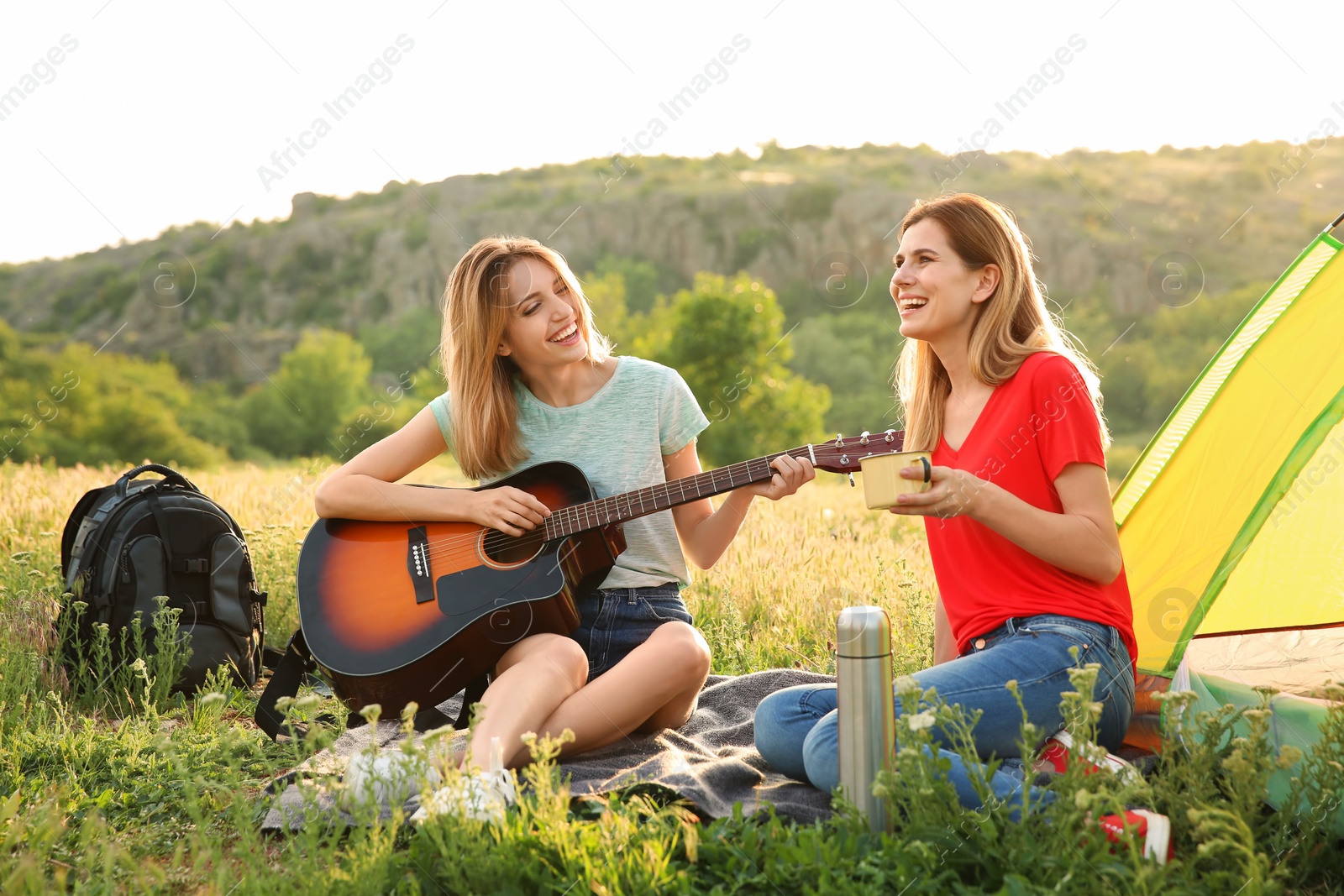 Photo of Young women resting with hot drink and guitar near camping tent in wilderness