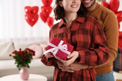 Happy couple celebrating Valentine's day. Beloved woman with gift box in room decorated with heart shaped air balloons, space for text