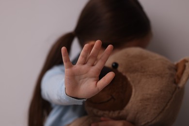Photo of Child abuse. Little girl with teddy bear doing stop gesture on light grey background, selective focus
