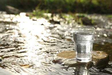 Photo of Glass of fresh water on stone near river outdoors. Space for text