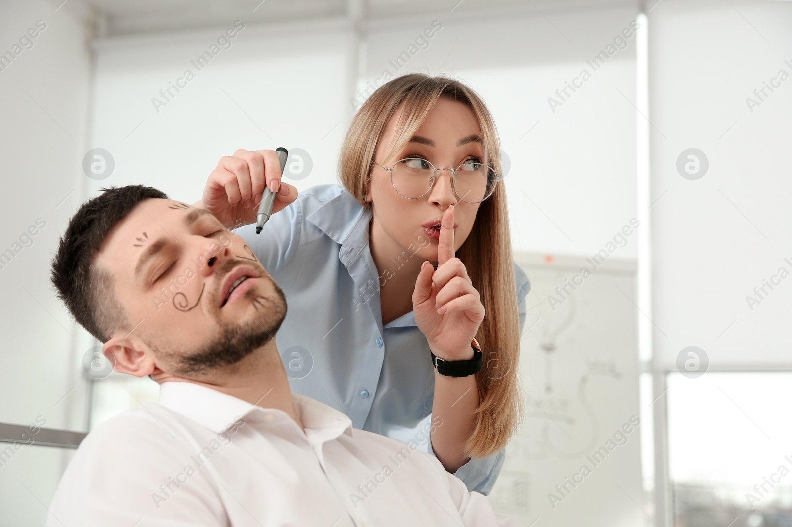 Photo of Young woman drawing on colleague's face while he sleeping in office. Funny joke