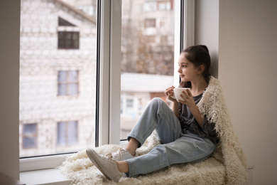 Photo of Cute little girl with cup of drink resting near window at home