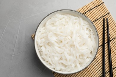 Photo of Bowl with cooked rice noodles, straw mat and chopsticks on light grey table, top view