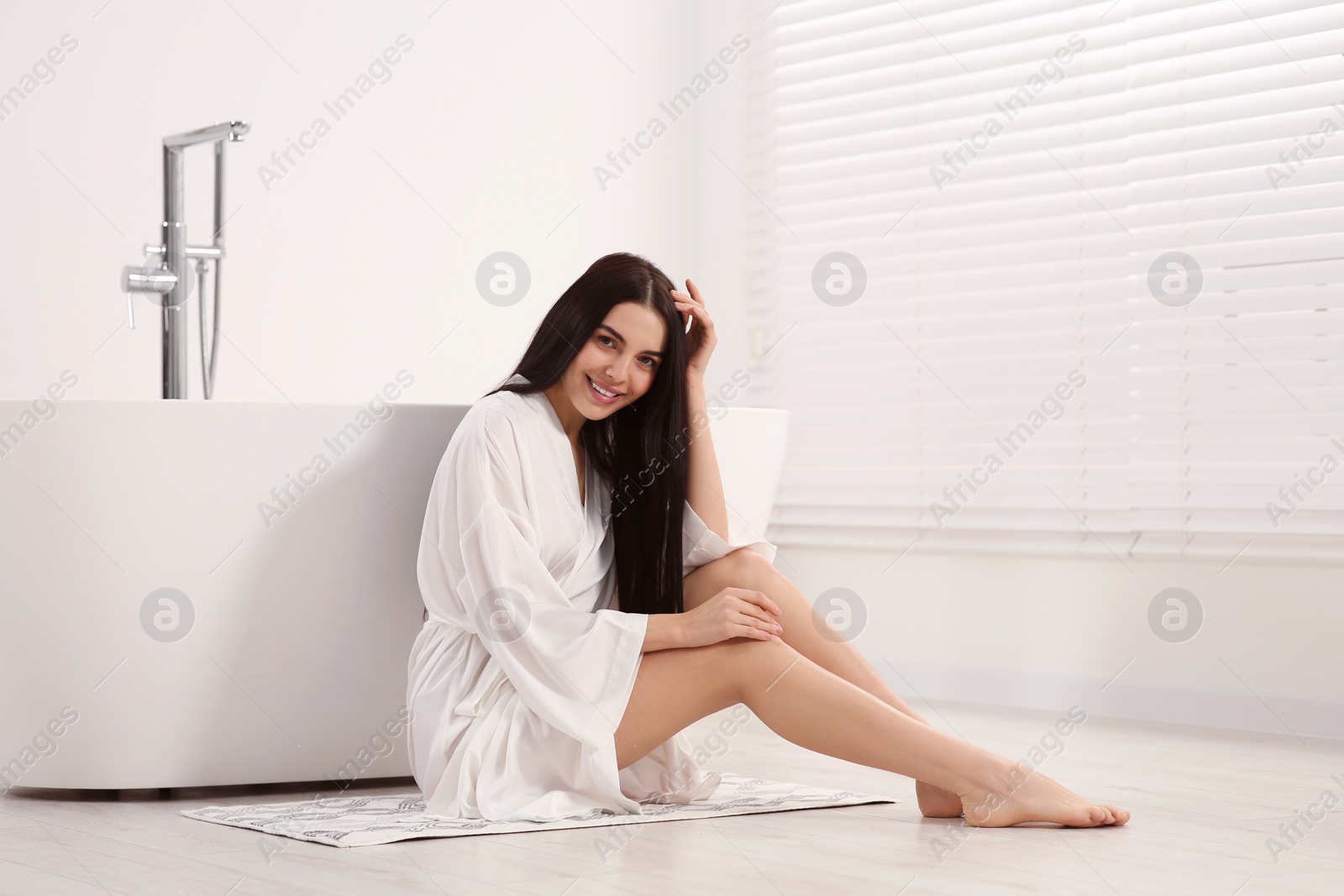 Photo of Beautiful happy woman wearing stylish bathrobe on floor near tub in bathroom