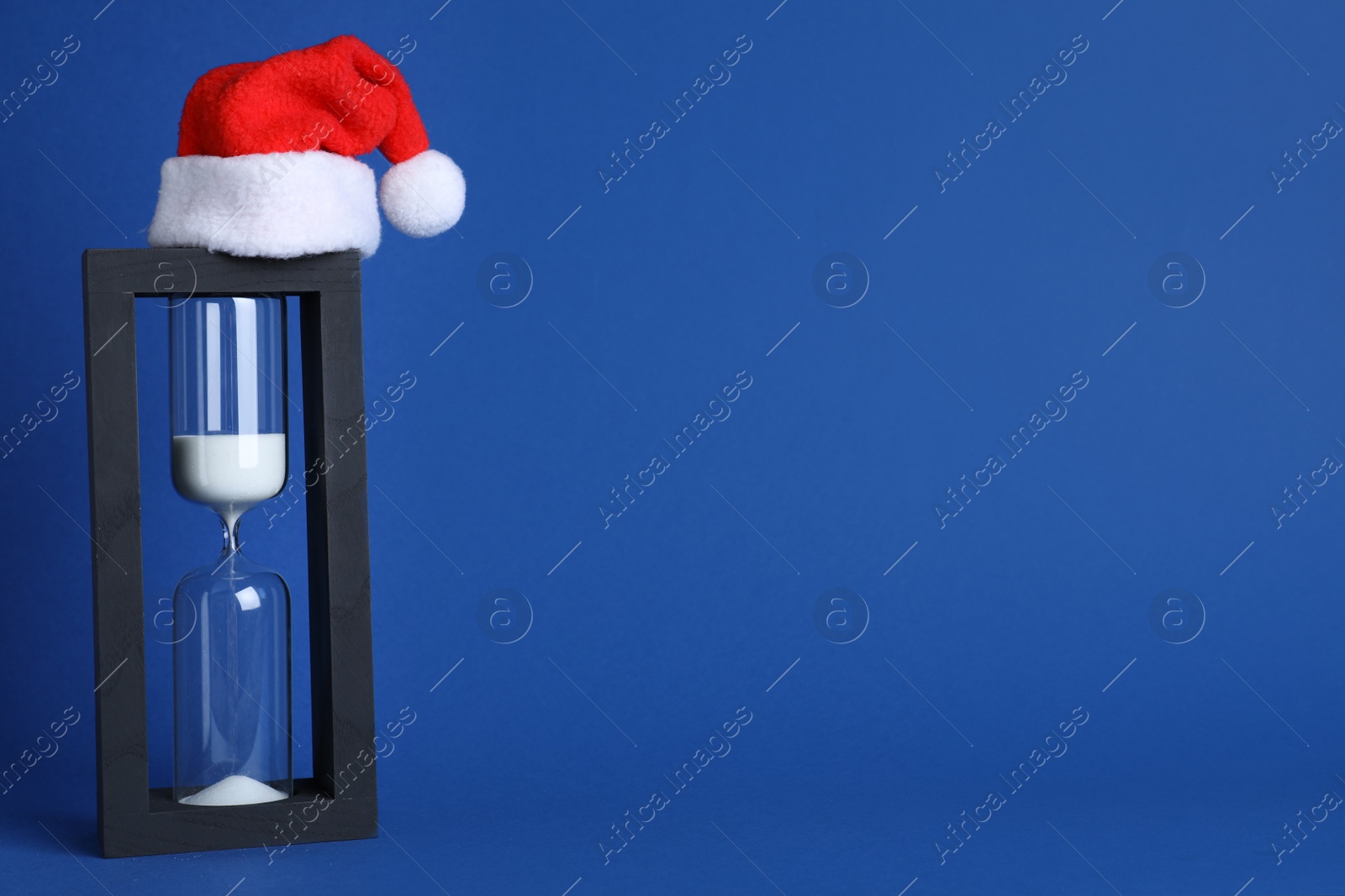 Photo of Hourglass and Santa hat on blue background, space for text. Christmas countdown