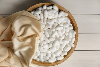 Photo of Cocoons with bowl and silk fabric on white wooden table, flat lay