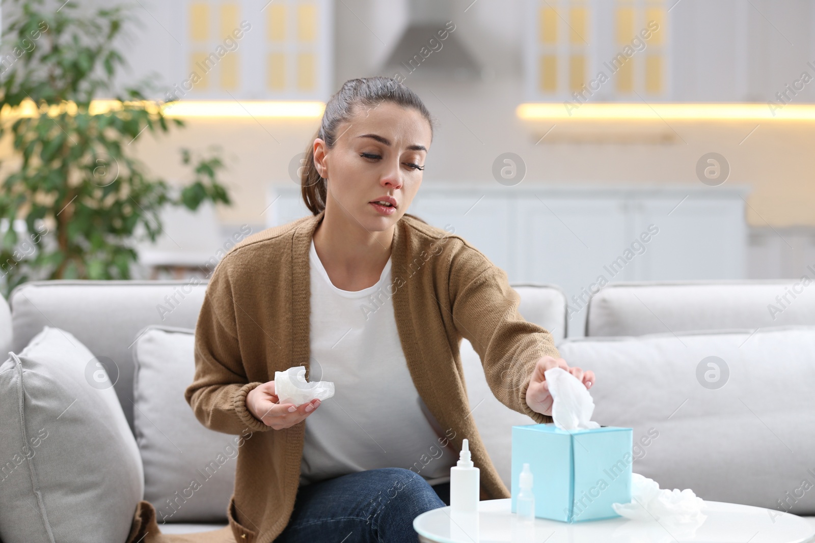 Photo of Sick woman with nasal spray, drops and box of paper tissues on sofa at home