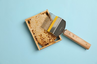 Photo of Hive frame with honeycomb and uncapping fork on light blue background, flat lay. Beekeeping