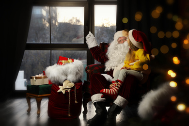 Photo of Santa Claus with little girl in armchair indoors. Christmas time