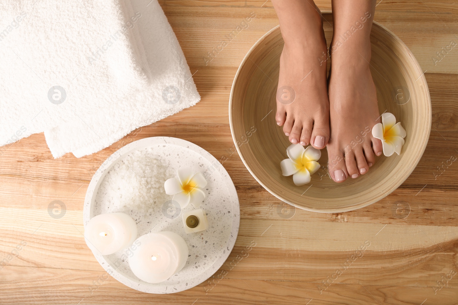 Photo of Woman soaking her feet in dish with water and flowers on wooden floor, top view. Spa treatment