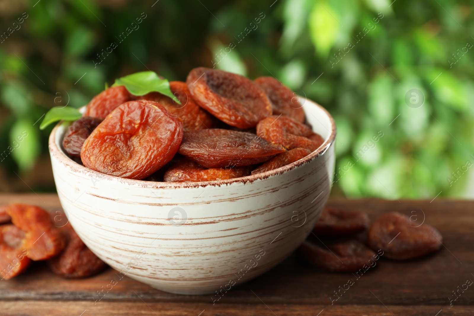 Photo of Bowl of tasty apricots on wooden table against blurred green background, space for text. Dried fruits