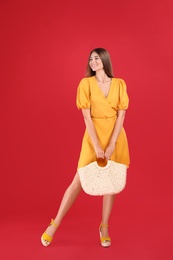 Photo of Young woman wearing stylish yellow dress with straw bag on red background