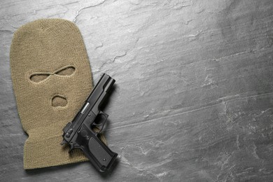 Beige knitted balaclava and pistol on black table, flat lay. Space for text