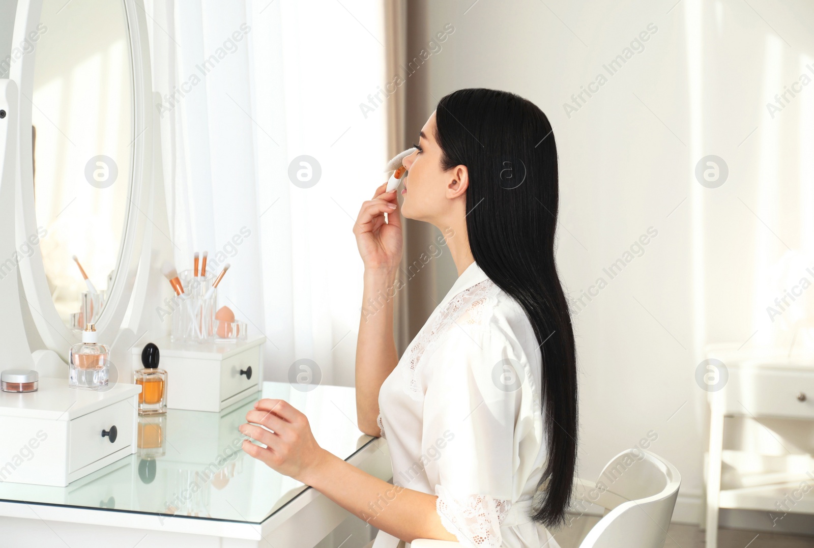 Photo of Beautiful young woman in robe doing make up at dressing table indoors