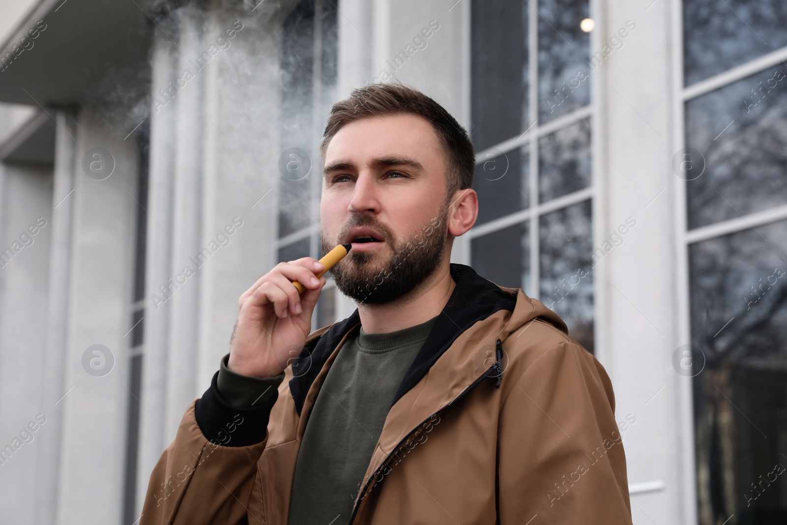 Photo of Handsome young man using disposable electronic cigarette outdoors