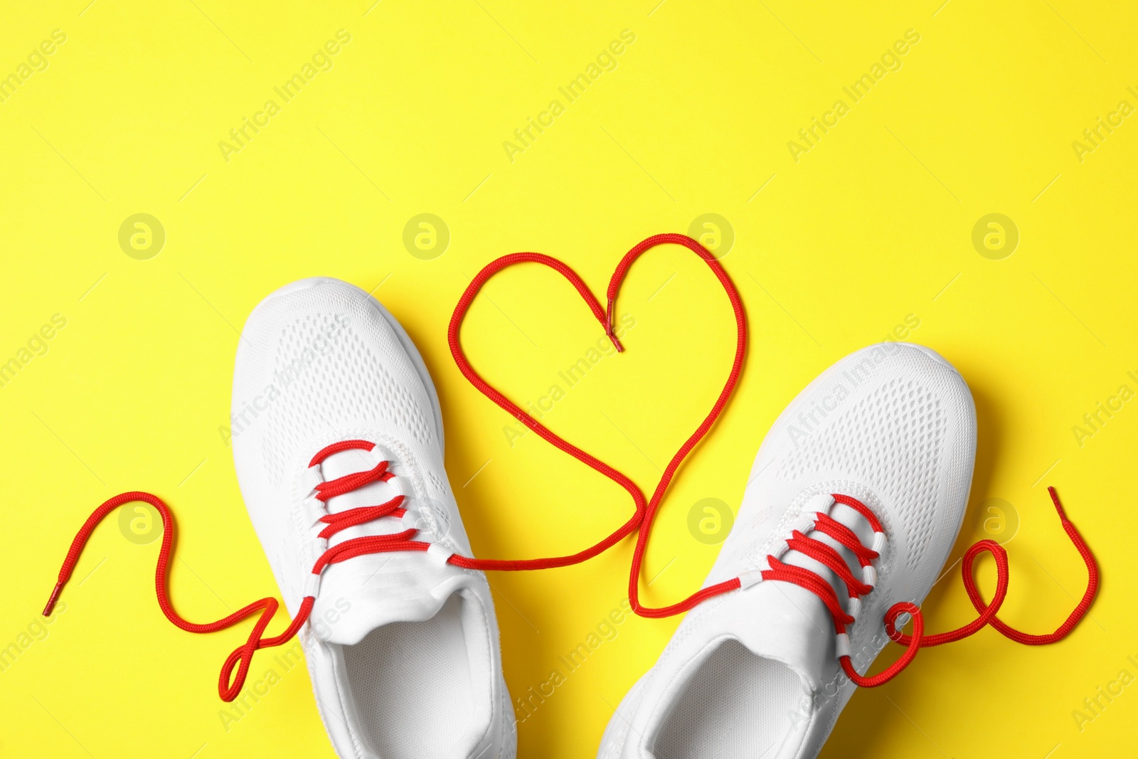 Photo of Stylish sneakers and red shoe laces in shape of heart on yellow background, flat lay