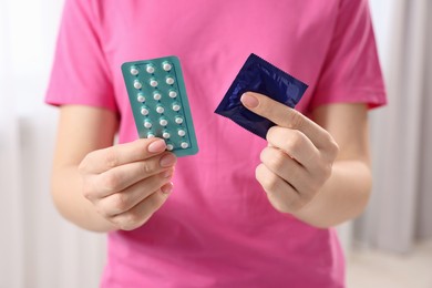 Woman holding condom and contraceptive pills on blurred background, closeup. Choosing birth control method
