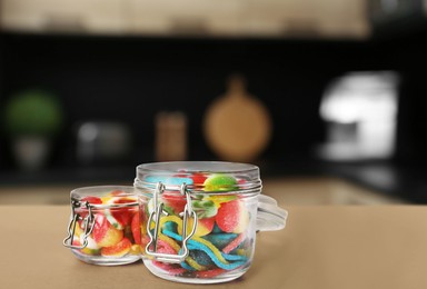 Glass jars with tasty gummy candies on light brown table in kitchen. Space for text