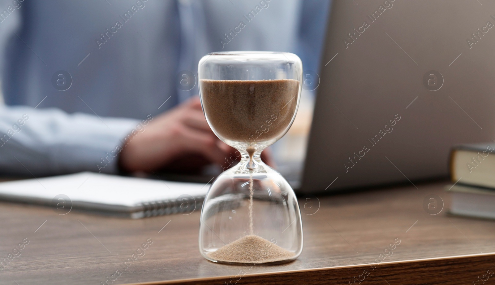 Photo of Hourglass with flowing sand on desk. Man using laptop indoors, selective focus