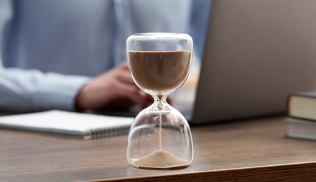 Hourglass with flowing sand on desk. Man using laptop indoors, selective focus