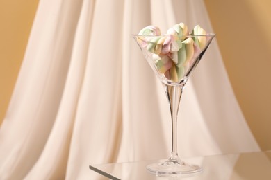 Photo of Martini glass with many tasty colorful marshmallows on table near white cloth. Space for text
