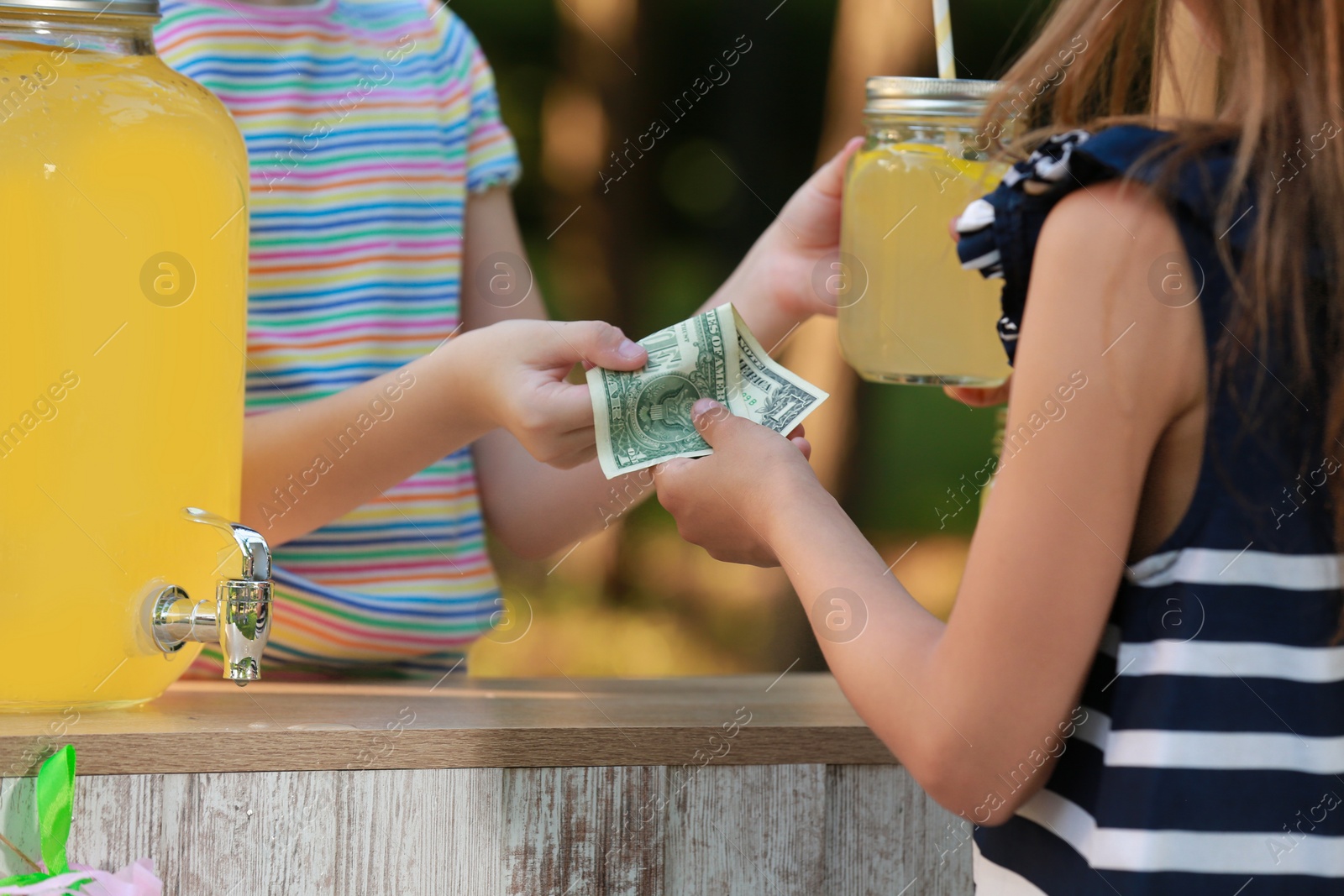 Photo of Little girl selling natural lemonade to kid in park, closeup. Summer refreshing drink