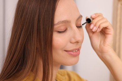 Photo of Beautiful woman applying serum onto eyelashes indoors, closeup