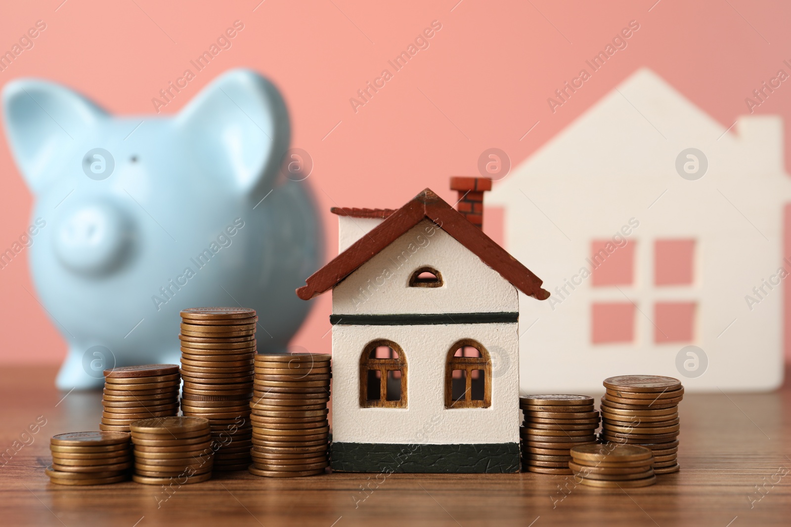 Photo of House models, piggy bank and stacked coins on wooden table against pink background, selective focus