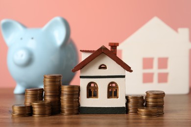 House models, piggy bank and stacked coins on wooden table against pink background, selective focus