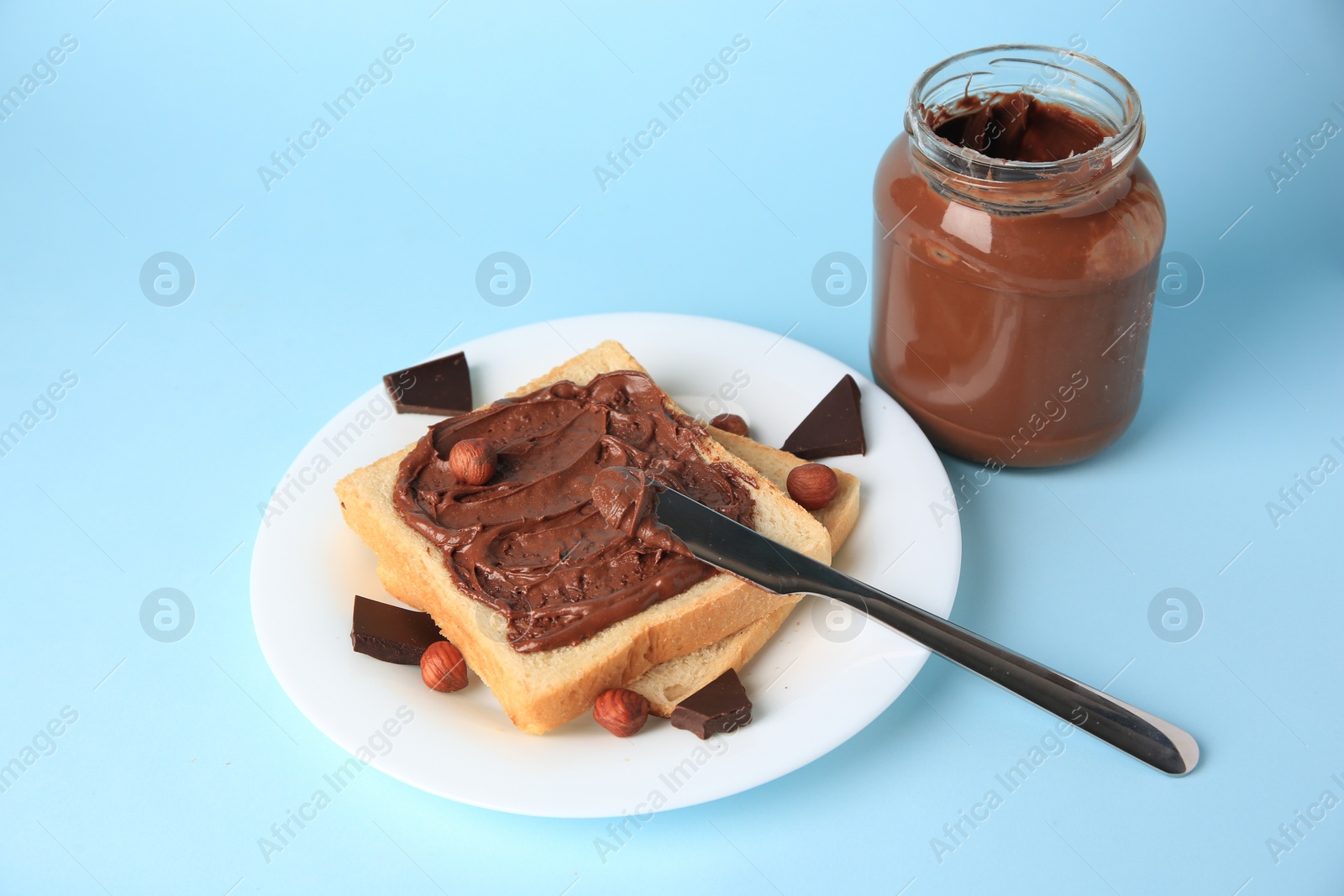 Photo of Tasty toast with chocolate paste and nuts near jar of cream on light blue background