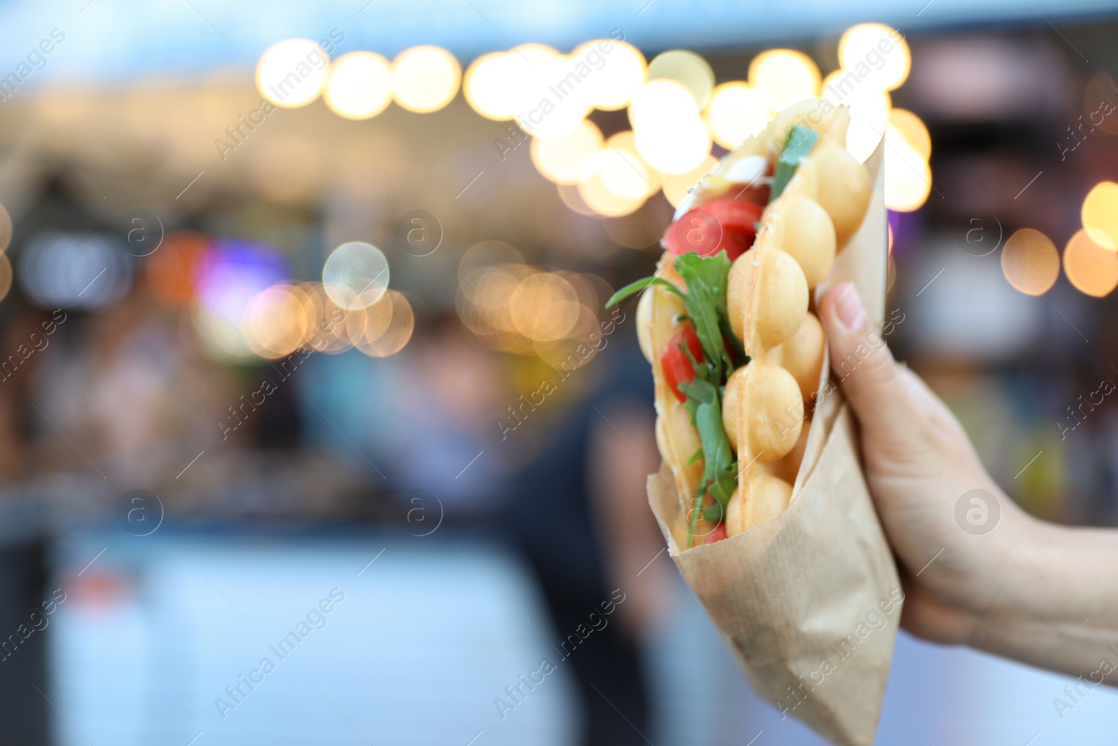 Photo of Young woman holding delicious bubble waffle with tomato and arugula outdoors, closeup