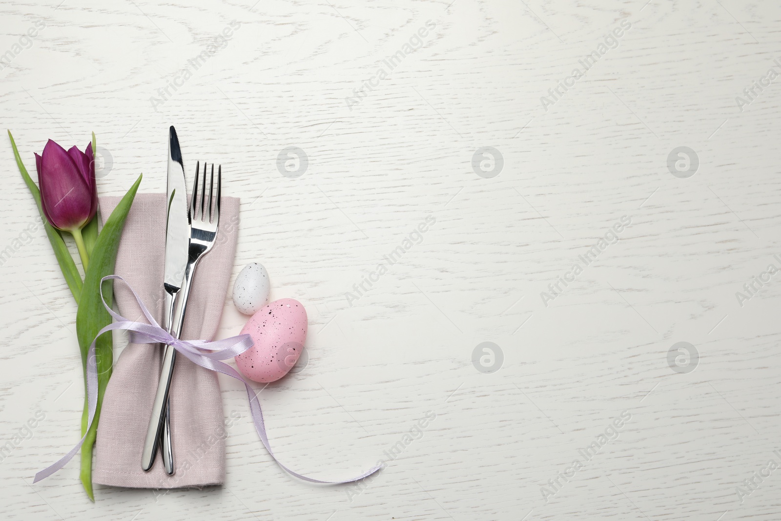 Photo of Cutlery set, painted eggs and beautiful flower on white wooden table, flat lay with space for text. Easter celebration