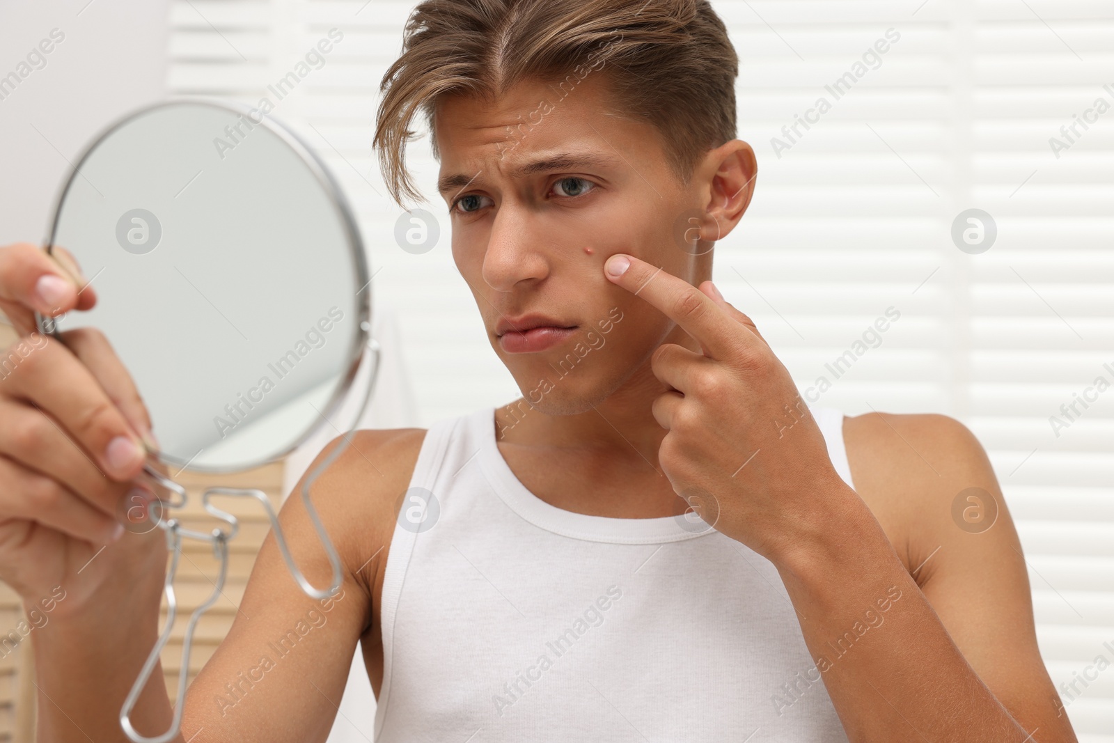 Photo of Upset young man looking at mirror and touching pimple on his face indoors. Acne problem