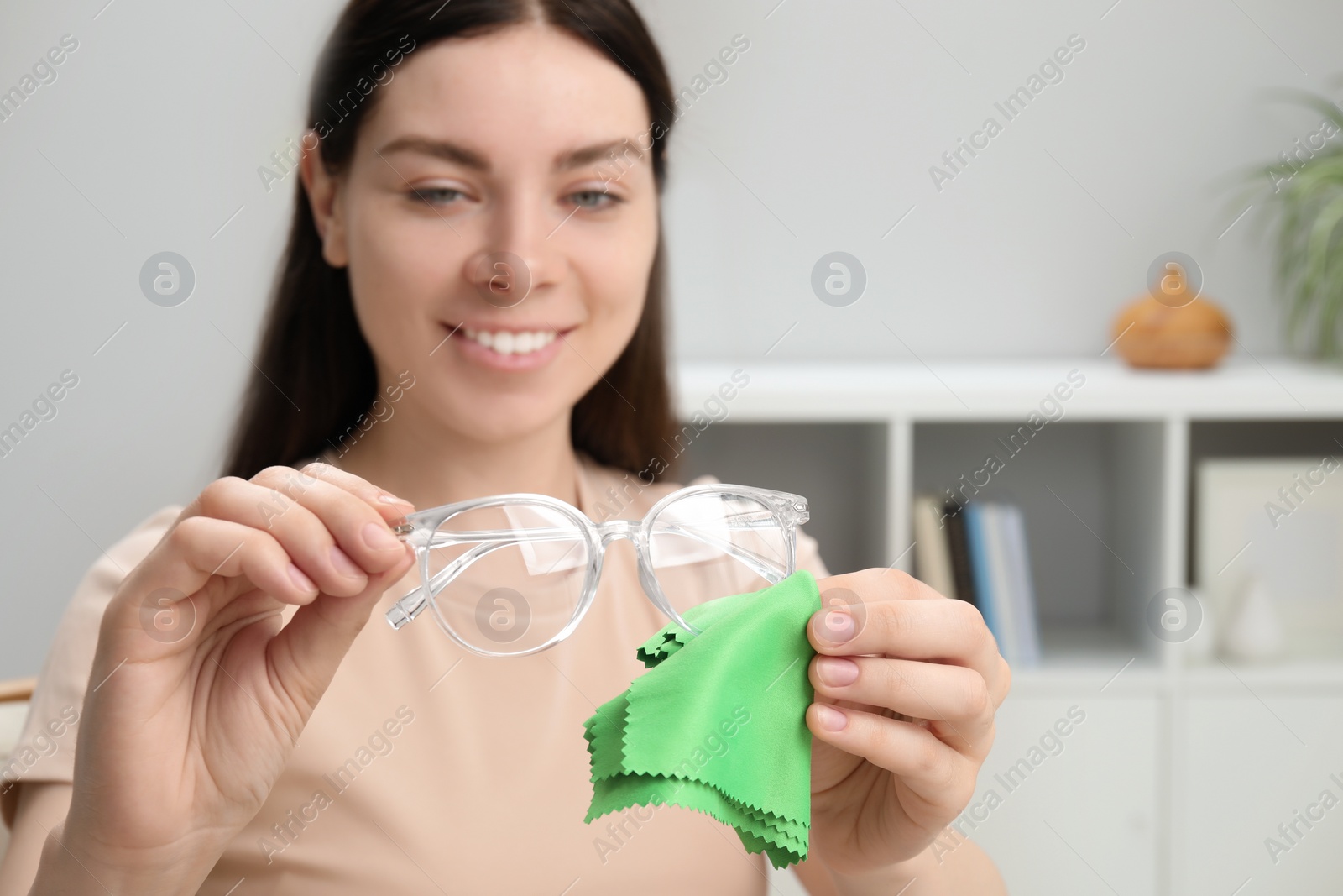 Photo of Beautiful woman cleaning glasses with microfiber cloth indoors, selective focus