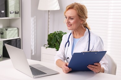 Photo of Doctor with laptop and clipboard at white table in clinic. Patient consultation