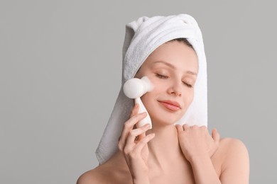 Young woman washing face with brush and cleansing foam on grey background. Space for text